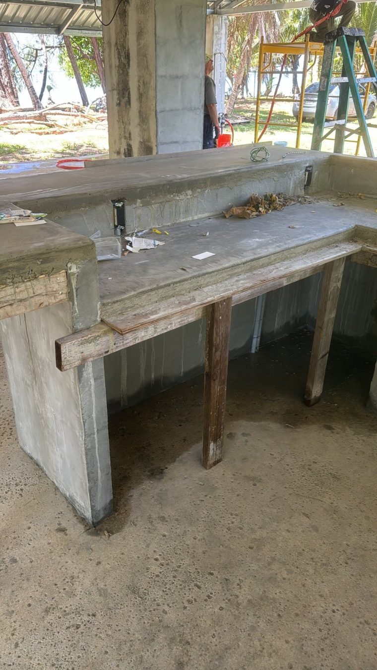 A poured concrete outdoor bar under a pavilion roof, facing the ocean through ironwood trees