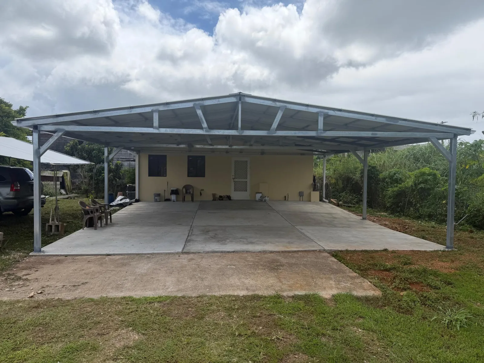 New galvanized steel roof framing and metal sheeting over a concrete slab beside a yellow house, heavy clouds above